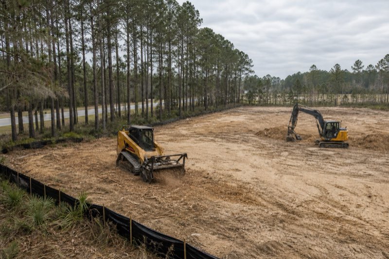 Excavator and tracked mulcher clearing a wooded commercial parcel beside a four-lane road, cleared pad area visible in the foreground